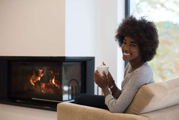 Felice gioiosa donna afroamericana che beve una tazza di caffè rilassante al camino. Giovane ragazza nera con riscaldamento di bevande calde in fase di riscaldamento. autunno a casa.