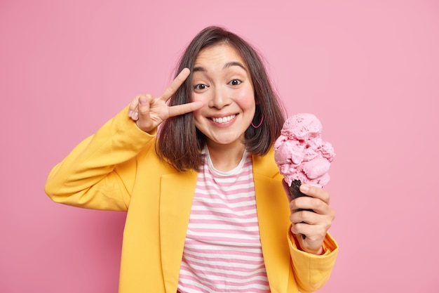 Felice gioiosa donna asiatica fa segno di vittoria sui sorrisi degli occhi tiene un grande cono gelato in cialda vestita con abiti eleganti si diverte durante la calda giornata estiva isolata sul muro rosa.