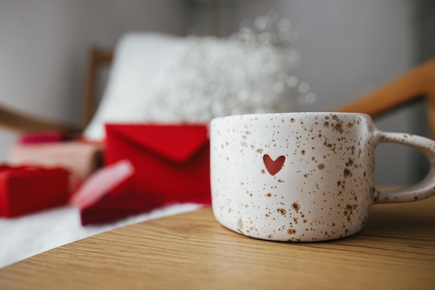 Felice giorno di San Valentino Simpatica tazza di tè moderna con cuore sul tavolo di legno contro una sedia elegante con regali busta rossa e fiori bianchi Sorpresa mattutina di San Valentino per l'amato