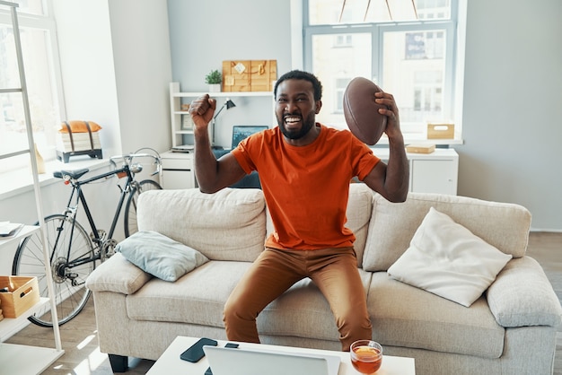 Felice giovane africano che applaude e sorride mentre guarda una partita sportiva a casa
