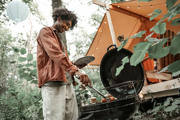 Felice giovane africano grigliare salsicce, fare barbecue per gli amici nella foresta in glamping. Tempo libero all'aperto. Riunione sociale dopo il lockdown. Bbq party, millennials che riposano fuori dal campeggio