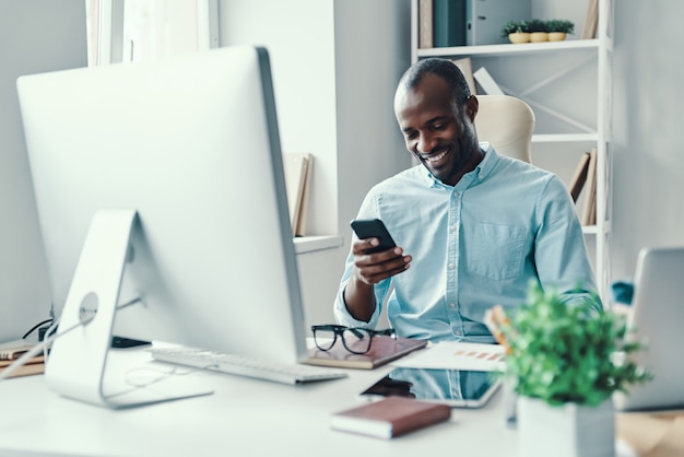 Felice giovane africano in camicia che usa lo smartphone e sorride mentre lavora in ufficio