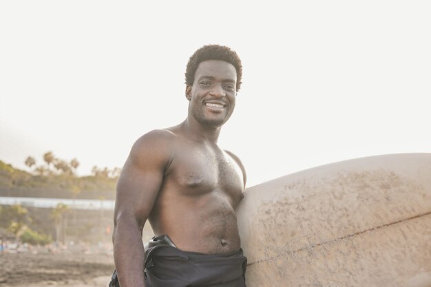 Felice giovane africano sorridente sulla fotocamera mentre tiene la tavola da surf sulla spiaggia Concetto di sport estremo