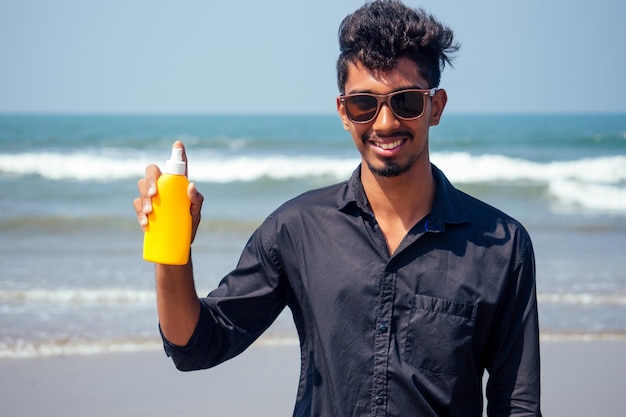 Felice giovane africano uomo sul modello beach.India maschio in possesso di una bottiglia di protezione solare studente adolescente sulla spiaggia di vacanza goa india.