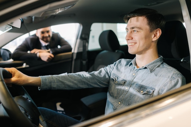 Felice giovane al volante di una nuova auto nella concessionaria di auto Venditore di auto professionista che indossa un tailleur racconta il nuovo modello di auto Concetto di scelta e acquisto di una nuova auto presso lo showroom