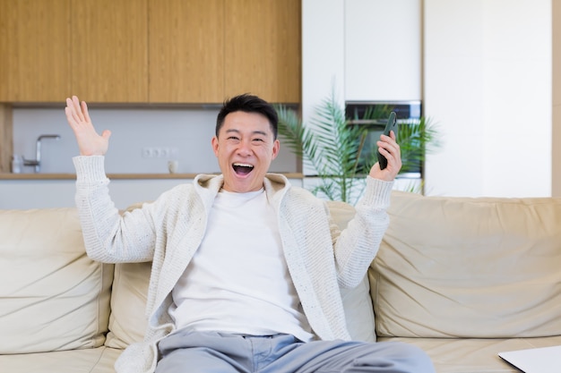 felice giovane asiatico a casa guardando il cellulare con emozione di vincitore