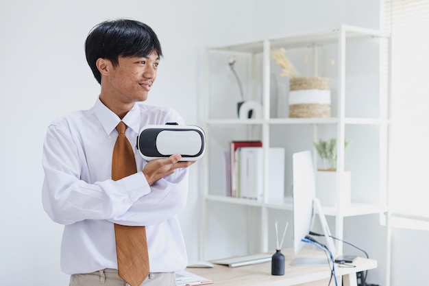 Felice giovane asiatico che indossa abiti formali tenendo gli occhiali VR a portata di mano e distogliendo lo sguardo Tecnologia