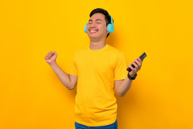 Felice giovane asiatico in maglietta casual che indossa le cuffie wireless tecnologiche per ascoltare musica tramite telefono cellulare isolato su sfondo giallo Concetto di stile di vita di emozioni della gente