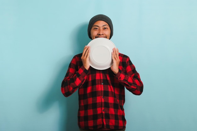 Felice giovane asiatico sorride mentre tiene un piatto bianco vuoto isolato su uno sfondo blu