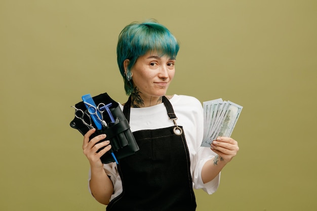 Felice giovane barbiere femminile che indossa l'uniforme che mostra la borsa da barbiere con strumenti da barbiere e denaro guardando la fotocamera isolata su sfondo verde oliva