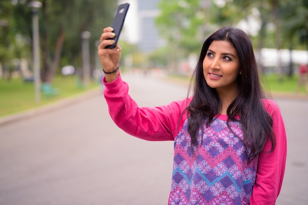 Felice giovane bella donna persiana prendendo selfie al parco