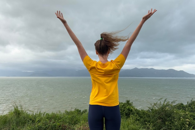 Felice giovane bella donna spensierata viaggiatore sta facendo un'escursione in viaggio godendosi la libertà camminando in cima alla montagna con vista sulla spiaggia del mare respirando in profondità l'aria dell'oceano del mare profondamente fresco alzando le mani