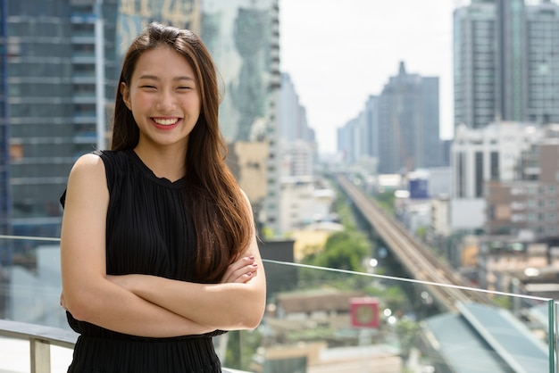 Felice giovane bella imprenditrice asiatica sorridente con le braccia incrociate contro la vista della città