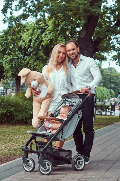 Felice giovane concetto di famiglia. Bella madre bionda e padre alla moda barbuto che camminano nel parco con la loro piccola figlia in un passeggino.
