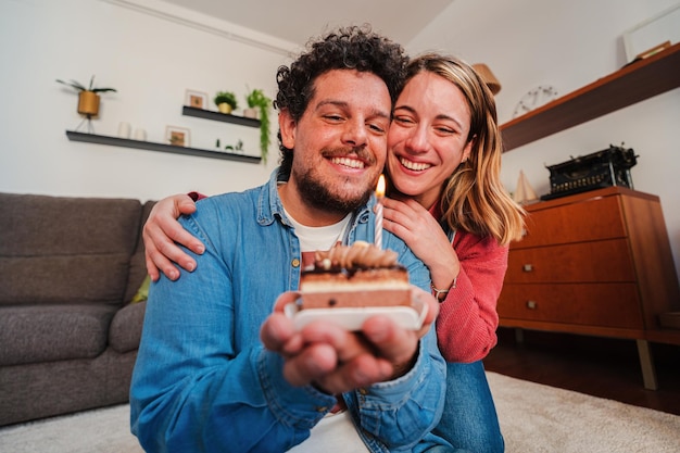 Felice giovane coppia che festeggia un compleanno con una piccola torta che soffia la candelina Moglie bionda che fa una divertente sorpresa al marito per il loro anniversario mentre lo abbraccia da dietro a casa