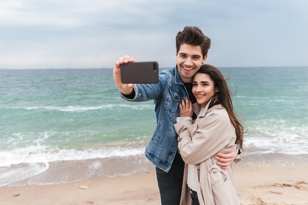 Felice giovane coppia che indossa cappotti autunnali trascorrono del tempo insieme al mare, facendo un selfie