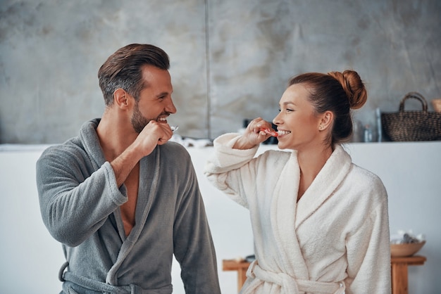 Felice giovane coppia in accappatoio che sorride e si pulisce i denti mentre fa la routine mattutina