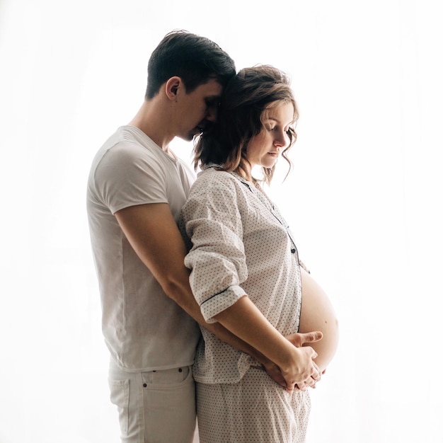 Felice giovane coppia incinta in pigiama bianco che abbraccia e tiene il pancione alla luce del mattino Elegante mamma e papà di famiglia incinta che si rilassano a casa in attesa del bambino La vera felicità