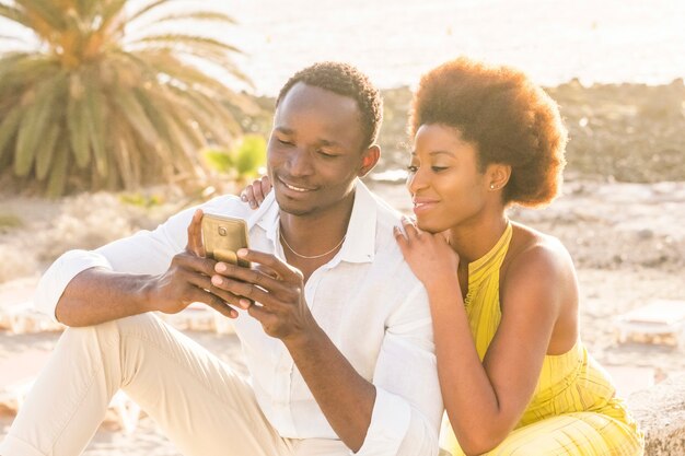 Felice giovane coppia nera in spiaggia con retroilluminazione del tramonto sullo sfondo, sorridere e ridere usando uno smartphone per chattare con gli amici o per vedere le foto della vacanza