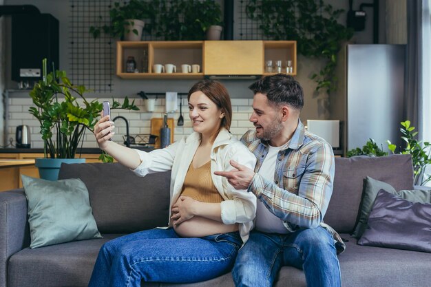 Felice giovane coppia sposata uomo e donna incinta utilizzando il telefono per la videochiamata abbracciando e seduto sul divano sorridendo e guardando lo schermo dello smartphone