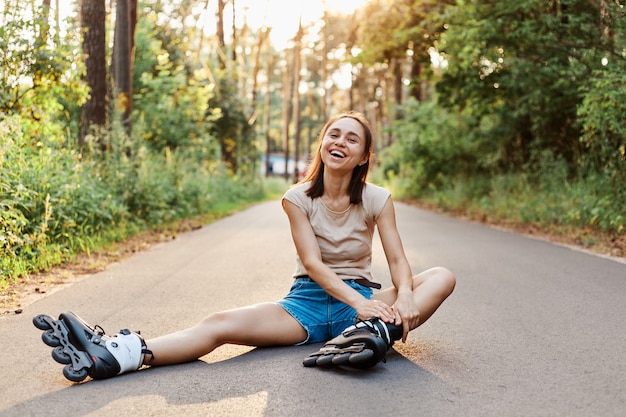 Felice giovane donna adulta in pattini, seduta sulla strada, indossando maglietta beige e jeans corti, guardando direttamente la telecamera e sorridendo felicemente, pattinaggio a rotelle femminile.