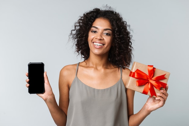 Felice giovane donna africana vestita casualmente in piedi isolato, tenendo il telefono cellulare con schermo vuoto, mostrando confezione regalo