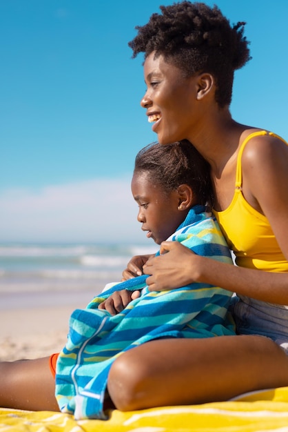 Felice giovane donna afroamericana seduta con la figlia avvolta in un asciugamano in spiaggia contro il cielo blu