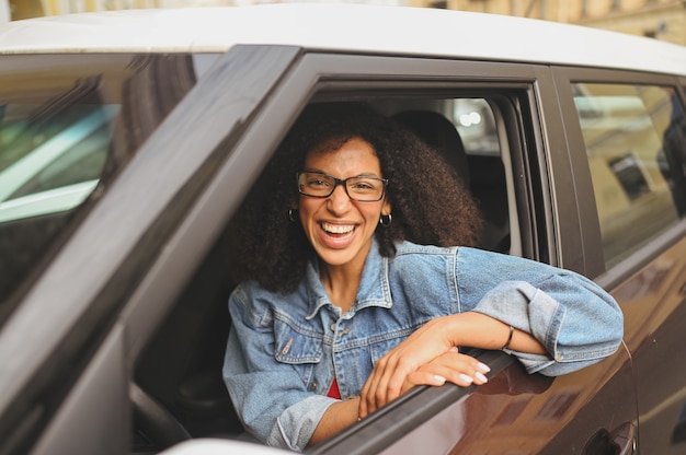 Felice giovane donna afroamericana sorridente autista dai capelli neri con gli occhiali seduto nella nuova auto marrone