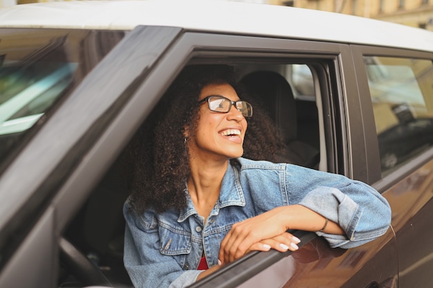 Felice giovane donna afroamericana sorridente autista dai capelli neri con gli occhiali seduto nella nuova auto marrone