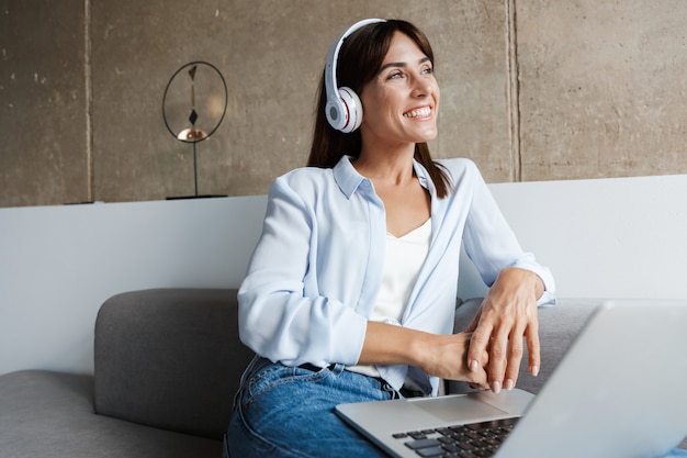 felice giovane donna al chiuso a casa utilizzando il computer portatile ascoltando musica con le cuffie sedersi sul divano in soggiorno.