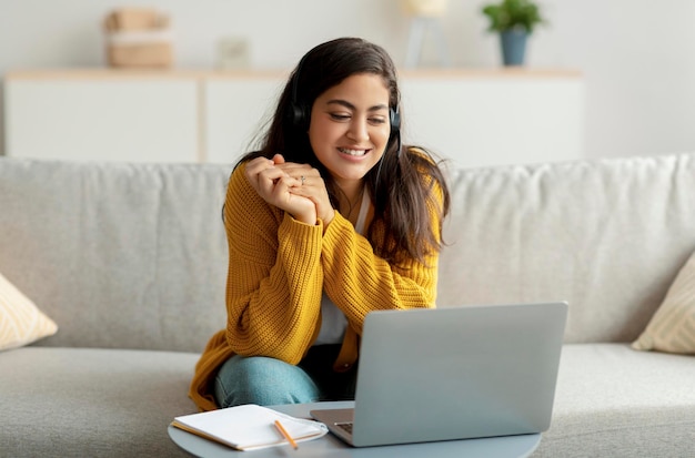 Felice giovane donna araba seduta sul divano che indossa l'auricolare e utilizza il laptop per guardare video o studiare a distanza da casa