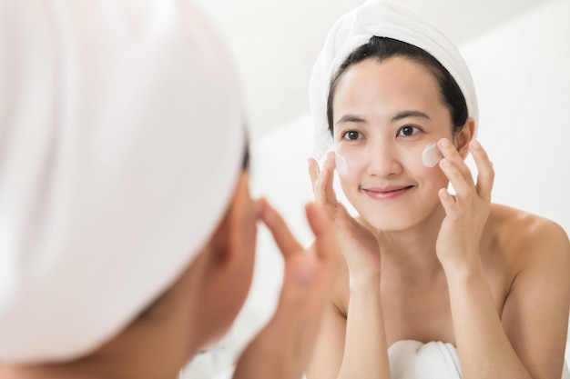 Felice giovane donna asiatica che applica lozioni per il viso mentre indossa un asciugamano e tocca il viso in bagno