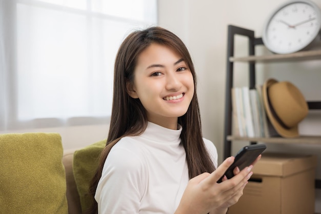 Felice giovane donna asiatica guardando lo schermo dello smartphone seduto sul divano in soggiorno che fa shopping online È molto felice Adolescente che legge le notizie dal telefono cellulare sentendosi eccitata