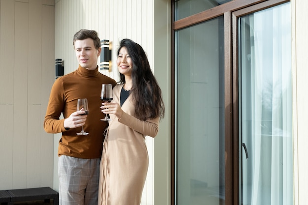 Felice giovane donna asiatica in elegante abito beige e suo marito con bicchieri di vino in piedi accanto alla porta nella camera d'albergo durante la celebrazione