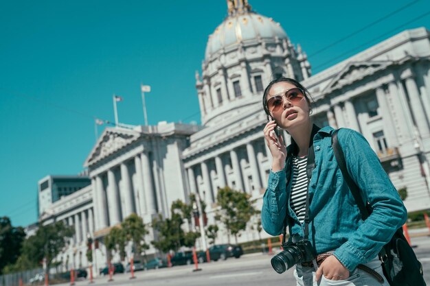 Felice giovane donna asiatica in occhiali da sole chiamando smartphone. famoso municipio di san francisco sullo sfondo sotto il cielo blu durante il giorno. ragazza turistica elegante con la macchina fotografica che parla sul cellulare.