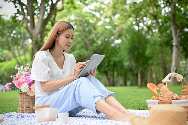 Felice giovane donna asiatica utilizzando tablet durante un picnic nel giardino verde in una giornata estiva