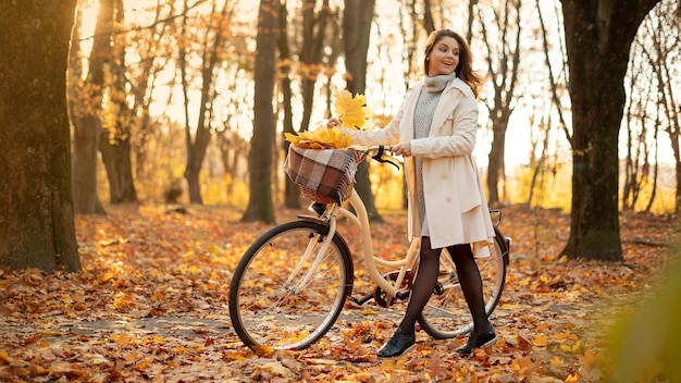 Felice giovane donna attiva con passeggiata in bicicletta d'epoca nel parco autunnale al tramonto Ragazza positiva con cesto pieno di foglie autunnali