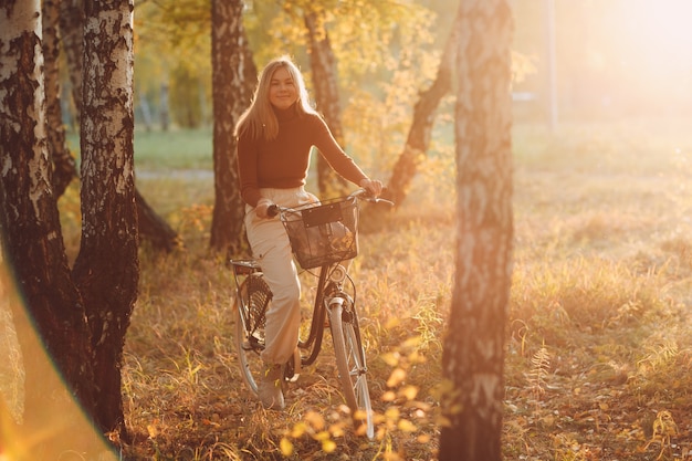 Felice giovane donna attiva in sella a una bicicletta d'epoca nel parco autunnale al tramonto.