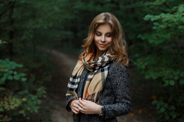 Felice giovane donna attraente in un cappotto grigio alla moda con un'elegante sciarpa a scacchi è in piedi su un sentiero nella foresta vicino agli alberi verdi