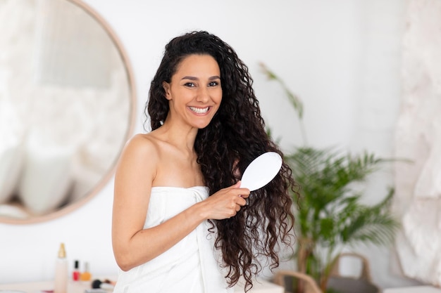 Felice giovane donna bruna caucasica in asciugamano che pettina lunghi capelli ricci con il pettine in camera da letto leggera