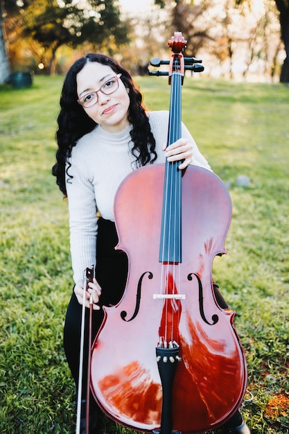 Felice giovane donna bruna con gli occhiali sorridente e suonare il violoncello al tramonto nel parco. Verticale.