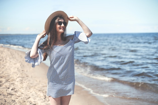 Felice giovane donna bruna in piedi sulla spiaggia dell'oceano mentre sorride e indossa cappello di moda, occhiali da sole. Il concetto di vacanza godendo.