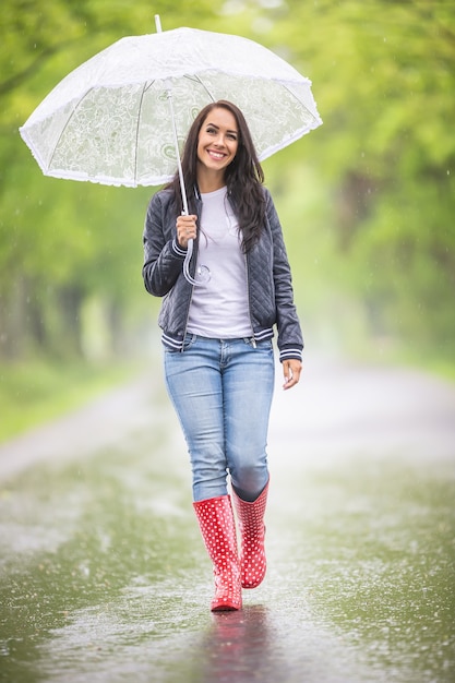 Felice giovane donna cammina con un ombrello sotto la pioggia nella natura indossando stivali da pioggia
