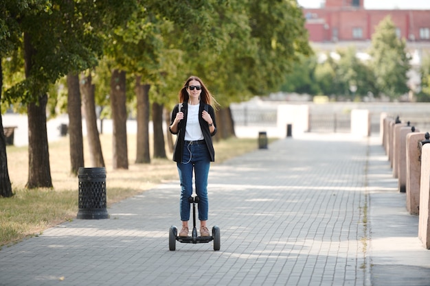 Felice giovane donna casual in occhiali da sole in movimento su scooter elettrico lungo la strada tra il fiume e la fila di alberi verdi al mattino