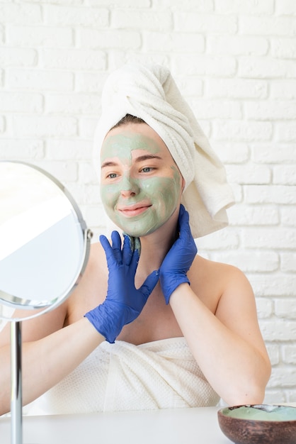 Felice giovane donna caucasica in asciugamani da bagno bianchi che indossano guanti applicando maschera di argilla guardando allo specchio