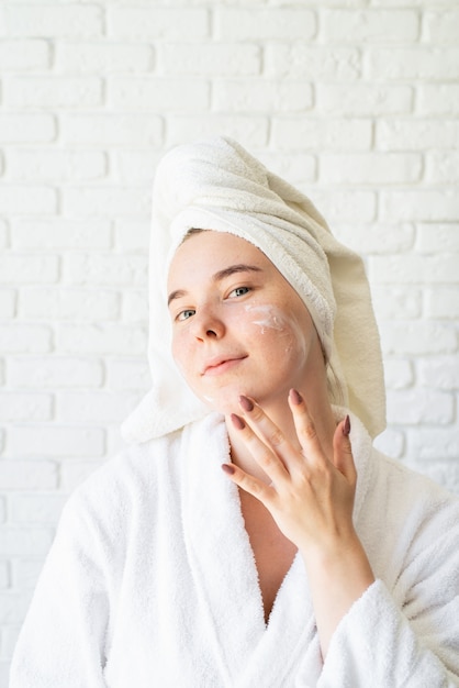 Felice giovane donna caucasica in telo da bagno bianco applicando la crema per il viso a casa