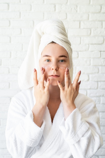 Felice giovane donna caucasica in telo da bagno bianco applicando la crema per il viso a casa