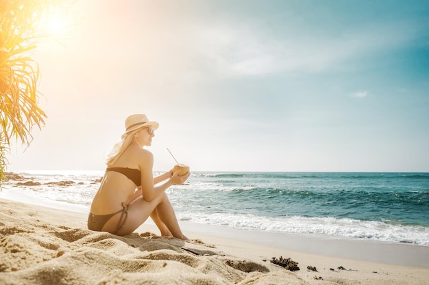 Felice giovane donna caucasica sorridente con il cappello con la noce di cocco nelle mani sulla spiaggia in un giorno d'estate soleggiato
