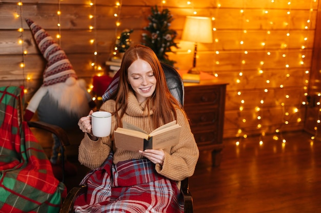 Felice giovane donna che beve caffè caldo e legge un libro mentre è seduto su una poltrona a dondolo con plaid