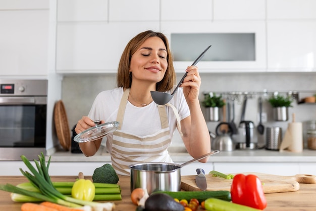 Felice giovane donna che cucina assaggiando la cena in una pentola in piedi in una cucina moderna a casa casalinga che prepara cibo sano sorridente casa e nutrizione ricette di dieta concetto